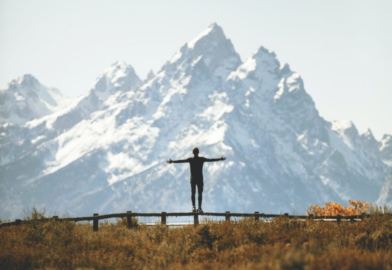 Individual standing on fence by mountains raising arms