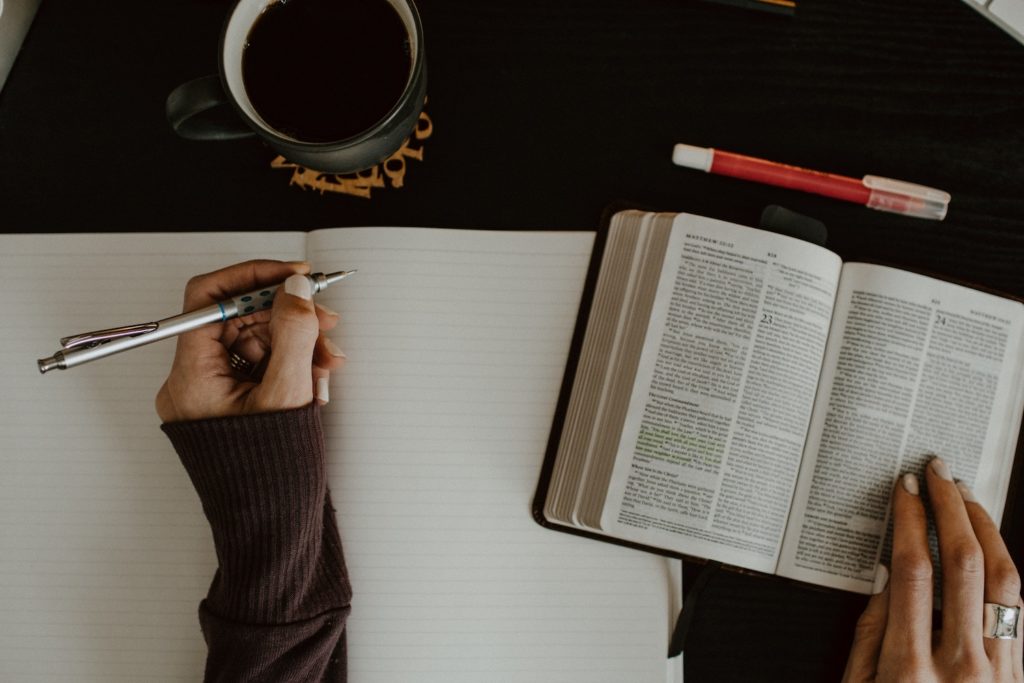 person writing in notebook with bible and coffee